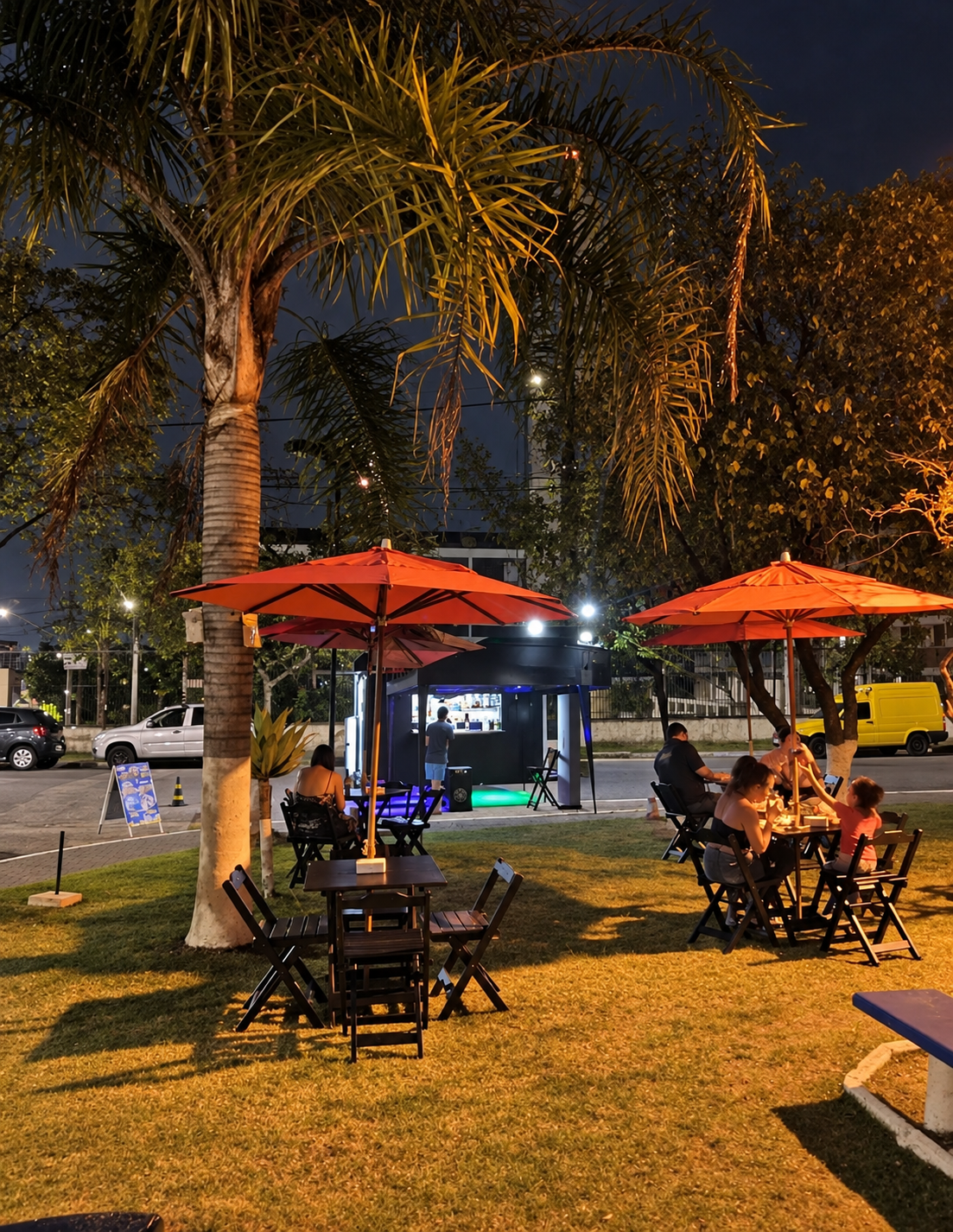 Espaço ao ar livre da Ferreira Burguer à noite, com mesas, ombrelones e palmeiras